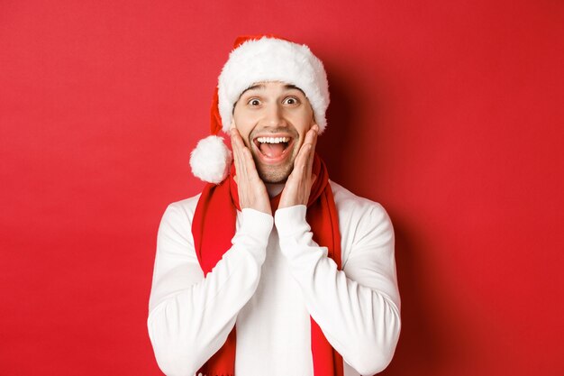 Concept of christmas, winter holidays and celebration. Close-up of surprised and happy man in santa hat and scarf, looking at something amazing, standing over red background.