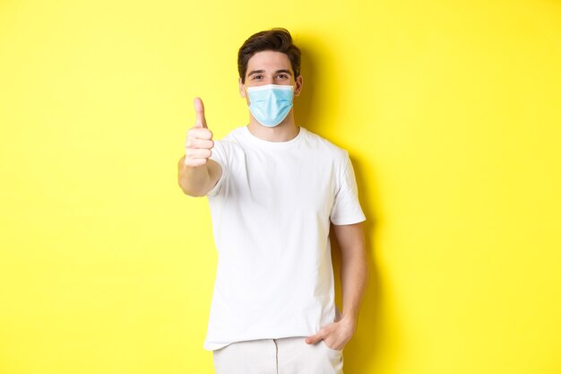 Concept of coronavirus, pandemic and social distancing. Confident young man in medical mask showing thumbs up, yellow background.