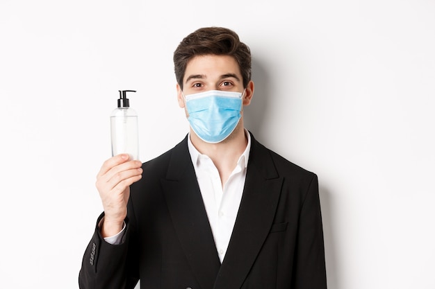 Concept of covid-19, business and social distancing. Close-up of handsome man in trendy suit and medical mask, showing hand sanitizer, standing against white background