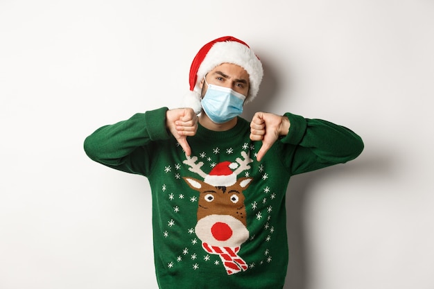 Free Photo concept of covid-19 and christmas holidays. displeased guy in face mask and santa hat showing thumbs down, standing over white background