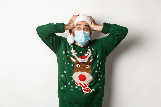 Free photo concept of covid-19 and christmas holidays. excited and super happy man in medical mask and santa hat looking amazed, white background