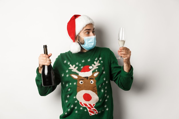 Concept of covid-19 and Christmas holidays. Man in medical mask and Santa hat enjoying glass of champagne, celebrating New Year, standing over white background