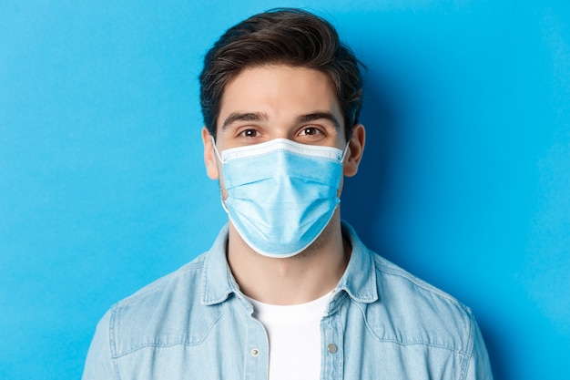 Concept of covid-19, pandemic and quarantine. Close-up of happy guy in medical mask looking at camera, standing over blue background.