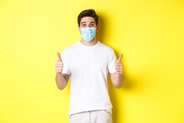 Concept of covid-19, quarantine and lifestyle. Happy man in medical mask showing thumbs up, approve or saying yes, like something good, yellow background.