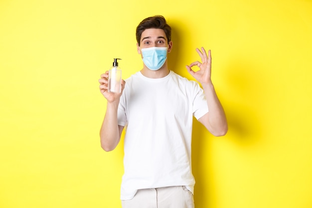 Concept of covid-19, quarantine and lifestyle. Satisfied young man in medical mask showing good hand sanitizer, make okay sign and recommending antiseptic, yellow background.