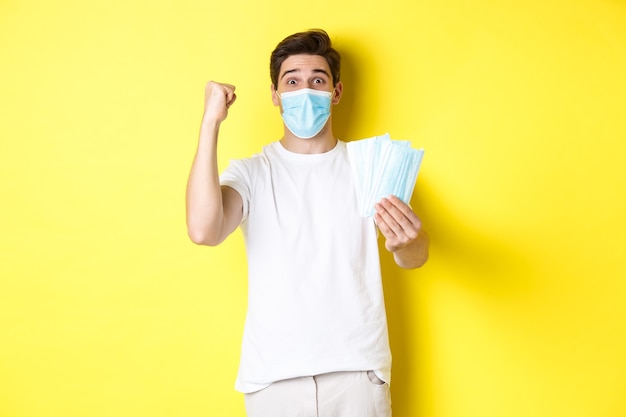 Concept of covid-19, quarantine and preventive measures. Happy man triumphing, raising hand to celebrate something and giving medical mask, standing against yellow background.