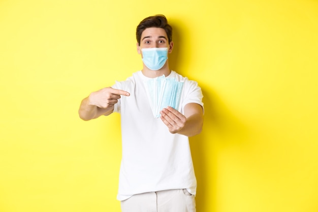 Concept of covid-19, quarantine and preventive measures. Young caucasian man giving medical masks for you, standing against yellow background