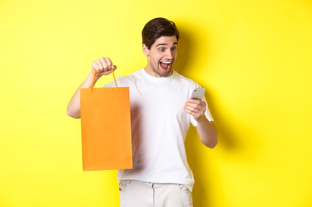 Concept of discounts, online banking and cashback. Surprised man showing shopping bag and looking happy at mobile screen, standing against yellow background.