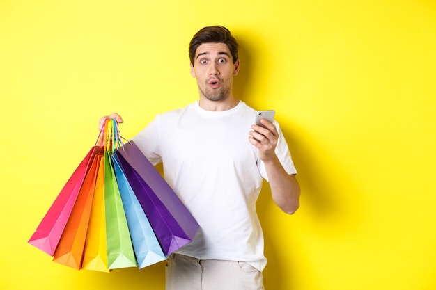 Concept of mobile banking and cashback. Surprised man holding shopping bags and smartphone, standing over yellow background.