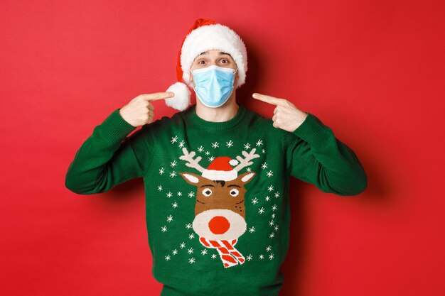 Concept of new year, coronavirus and social distancing. Happy man in santa hat and christmas sweater, recommending to wear medical mask on party, standing over red background