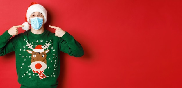 Free Photo concept of new year, coronavirus and social distancing. happy man in santa hat and christmas sweater, recommending to wear medical mask on party, standing over red background.