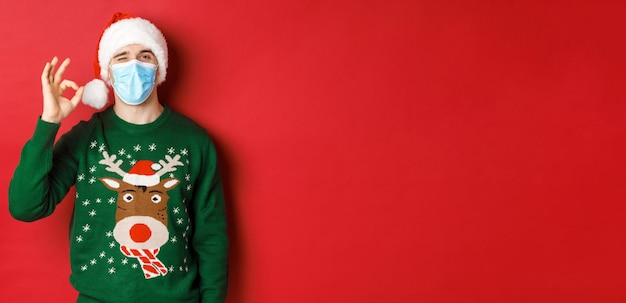 Free Photo concept of new year, covid-19 and social distancing. cheerful man in medical mask and santa hat, showing okay sign, recommending something good, standing over red background.