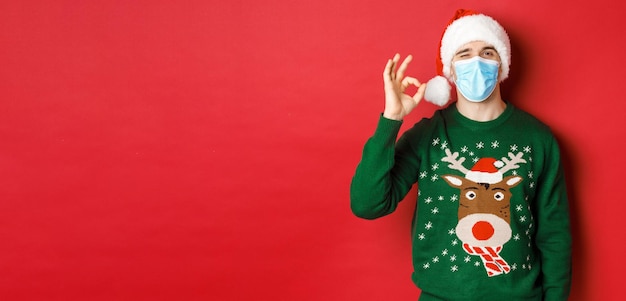 Free Photo concept of new year covid and social distancing cheerful man in medical mask and santa hat showing o...