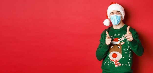 Concept of new year covid19 and social distancing Cheerful man in christmas sweater medical mask and santa hat pointing fingers at camera wishing happy holidays standing over red background