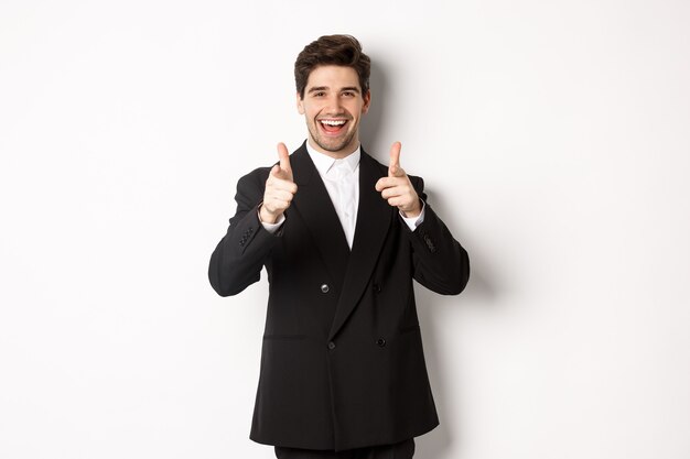 Concept of new year party, celebration and lifestyle. Handsome and successful guy in black suit, pointing fingers at camera and congratulating you, standing over white background.