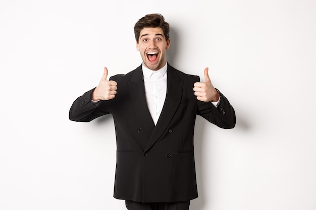 Free photo concept of new year party, celebration and lifestyle. portrait of amazed and pleased handsome man in black suit, showing thumbs-up, like product, approve something good, white background