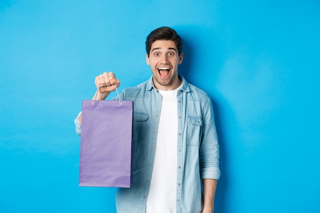 Concept of shopping, holidays and lifestyle. Excited handsome guy holding paper bag with gift and looking happy, standing over blue background