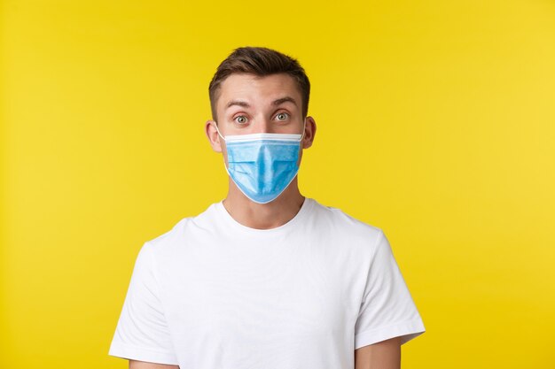 Concept of social distancing, covid-19 and people emotions. Excited and surprised handsome guy found out awesome news, wearing medical mask, looking amazed over yellow background.