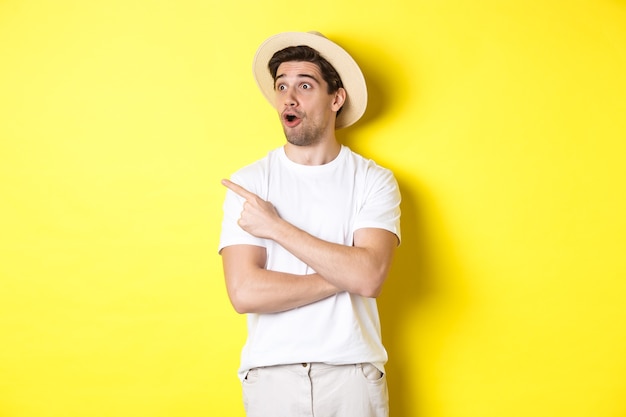 Free photo concept of tourism and lifestyle. excited handsome guy in straw hat checking out advertisement, pointing and looking at upper left corner logo, yellow background