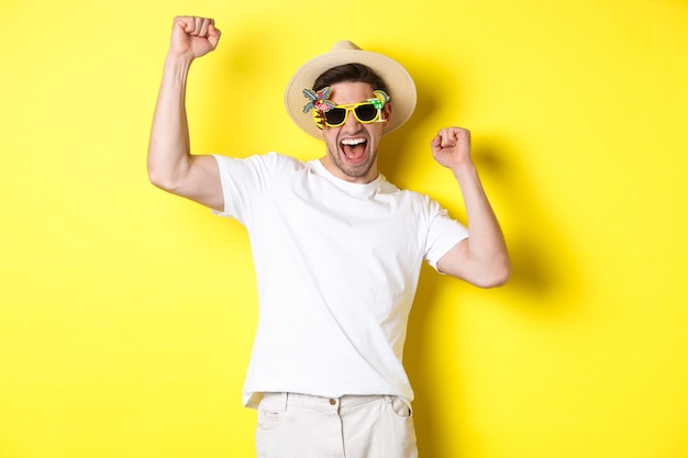 Free photo concept of tourism and lifestyle. happy lucky guy winning trip, rejoicing and wearing holiday outfit, summer hat and sunglasses, yellow background.