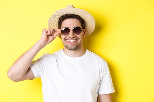 Concept of tourism and vacation. Close-up of handsome man tourist looking happy, wearing sunglasses and summer hat, standing over yellow background