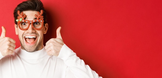 Free photo concept of winter holidays, christmas and celebration. close-up of excited handsome man in party glasses, smiling and showing thumbs-up in approval, standing over red background.