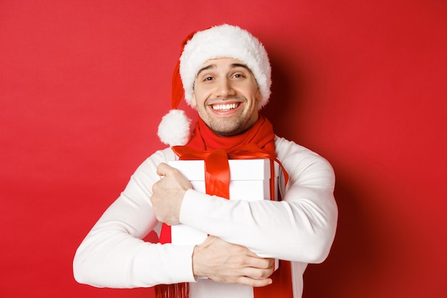 Concept of winter holidays, christmas and lifestyle. Image of lovely guy in santa hat and scarf, hugging his new year present and smiling flattered, standing over red background.