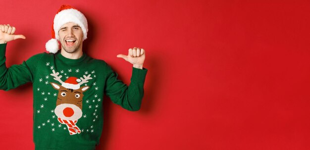 Concept of winter holidays christmas and lifestyle Sassy handsome man in santa hat and green sweater pointing at himself and winking standing over red background