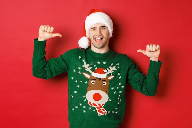 Free photo concept of winter holidays, christmas and lifestyle. sassy handsome man in santa hat and green sweater, pointing at himself and winking, standing over red background