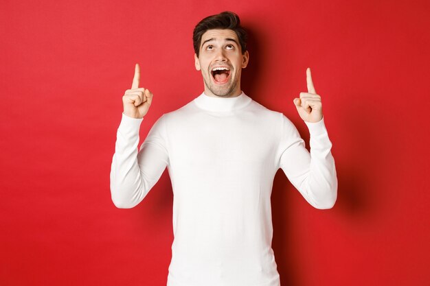 Concept of winter holidays. Image of amazed handsome guy reacting to christmas announcement, pointing and looking up at copy space, standing over red background