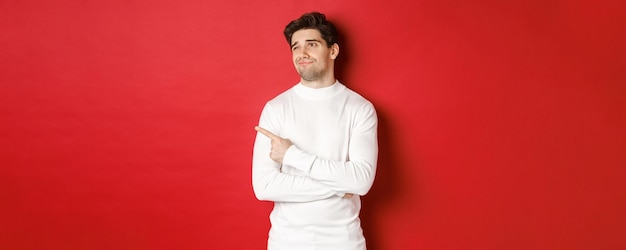 Concept of winter holidays image of displeased young man in white sweater looking and pointing finge