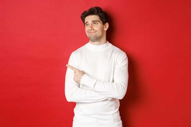 Concept of winter holidays. Image of displeased young man in white sweater, looking and pointing finger left at something average, standing skeptical over red background