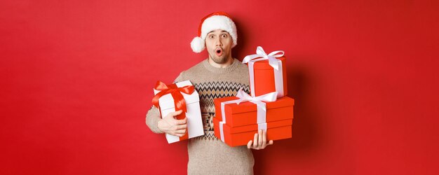 Concept of winter holidays, new year and celebration. Image of surprised handsome guy in sweater and santa hat, receiving gifts, holding boxes with presents and looking amazed.