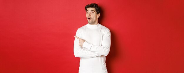 Concept of winter holidays portrait of impressed handsome man in white sweater pointing and looking