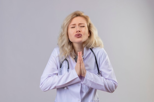 Free photo concered young doctor wearing stethoscope in medical gown showing pray gesture on isolation white wall