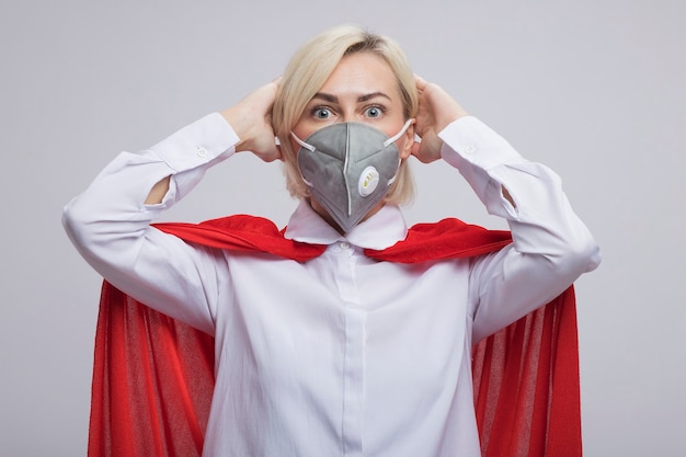 Free photo concerned middle-aged blonde superhero woman in red cape wearing protective mask  putting hands on head isolated on white wall