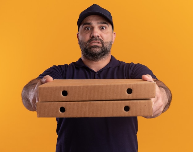 Free photo concerned middle-aged delivery man in uniform and cap holding out pizza boxes at front isolated on yellow wall