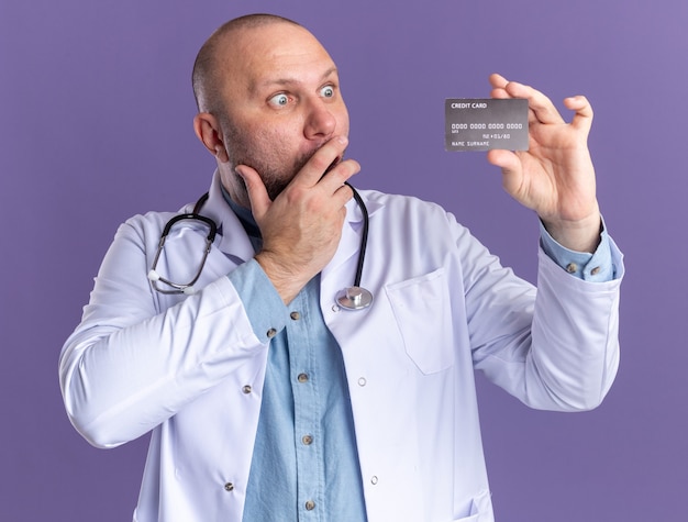 Concerned middle-aged male doctor wearing medical robe and stethoscope holding and looking at credit card keeping hand on mouth isolated on purple wall