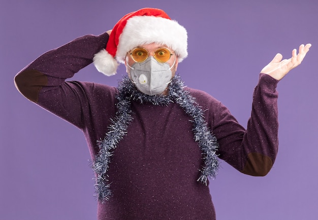 Free Photo concerned middle-aged man wearing santa hat and tinsel garland around neck with glasses and protective mask looking at camera showing empty hand keeping hand behind head isolated on purple background