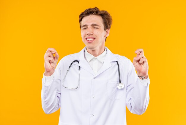 Concerned with closed eyes young male doctor wearing medical robe with stethoscope crossing fingers