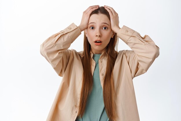 Concerned and worried girl panicking holding hands on head with opened mouth looking troubled having big problem standing confused against white background