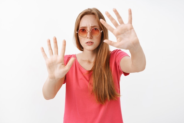 Concerned and worried young redhead female in sunglasses and pink t-shirt starting get nervous opening mouth frowning raising palms near chest as if being stuck in box, restraining from dangerous man