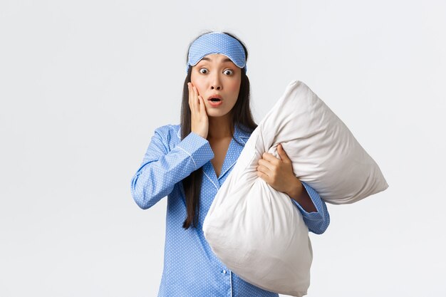 Concerned and worried young woman in blue pajamas and sleeping mask, holding pillow and gasping stare anxious, feel compassion or pity for someone got in trouble, hear shocking news.