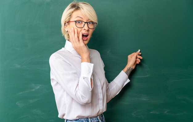 concerned young blonde female teacher wearing glasses in classroom standing in profile view in front of chalkboard pointing at chalkboard with chalk looking at front keeping hand on face