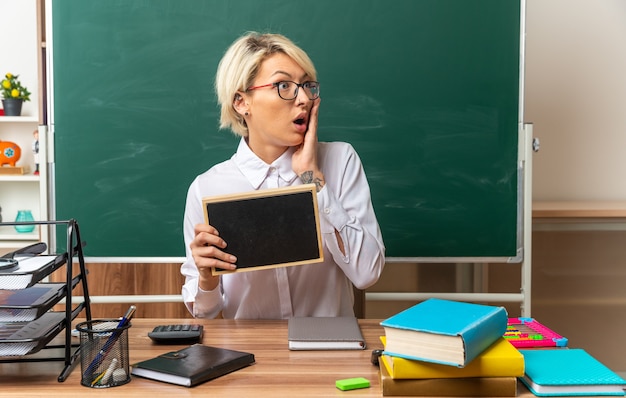 concerned young blonde female teacher wearing glasses sitting at desk with school supplies in classroom showing mini blackboard keeping hand on face looking at side