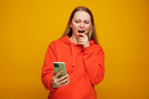 Concerned young blonde woman holding and looking at mobile phone keeping hand on chin 