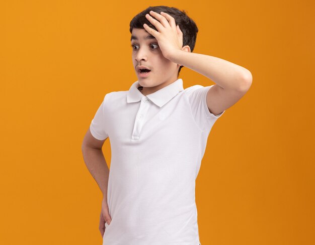 Concerned young boy putting hand on head keeping another one on waist looking at side isolated on orange wall