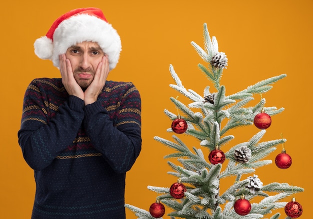 Free Photo concerned young caucasian man wearing christmas hat standing near decorated christmas tree keeping hands on face  isolated on orange wall