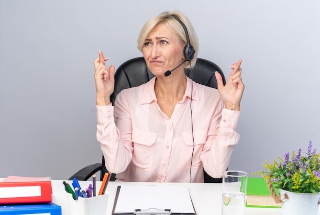 Concerned young female call center operator wearing headset sitting at table with office tools crossing fingers 