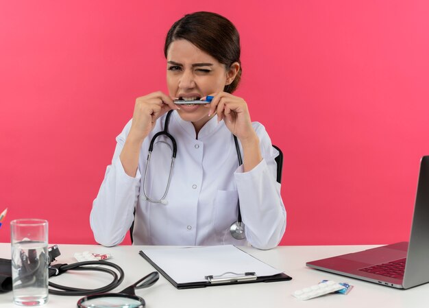 Concerned young female doctor wearing medical robe with stethoscope sitting at desk work on computer with medical tools holding pen in mouth on isolated pink wall with copy space